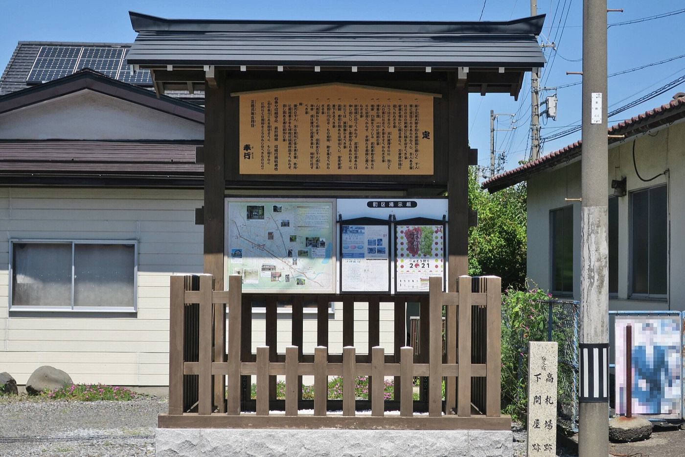 中山道塩尻宿の高札場跡に復元された高札場。屋根を載せた柱に「定」で始まる大きな札が掲げられ、その下に「町区掲示板」があって地図や催しのポスターが貼られている。手前に木の柵、右手に「高札場跡 下問屋跡」の石柱